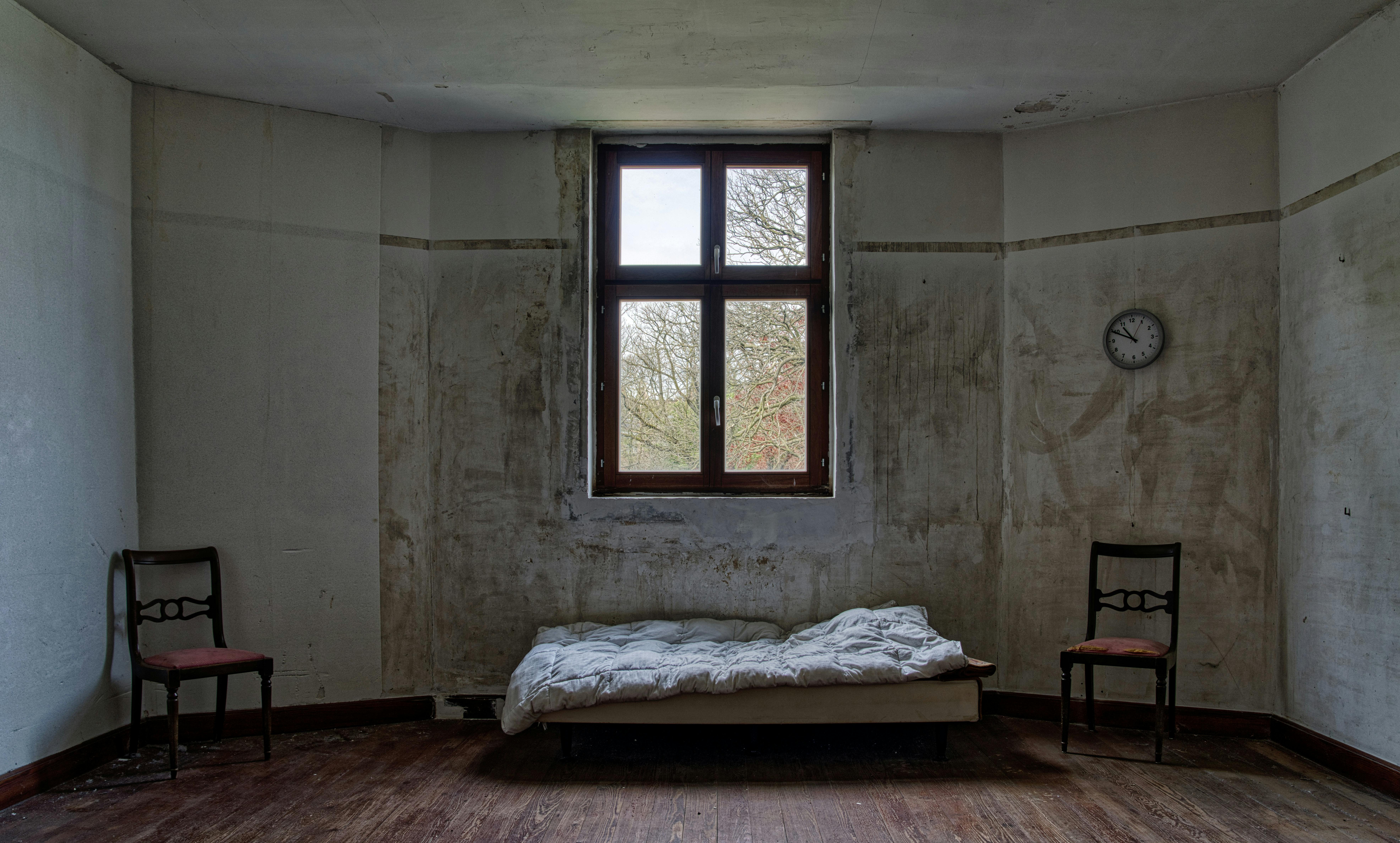 An old, abandoned room with a single bed, two chairs, and a window.