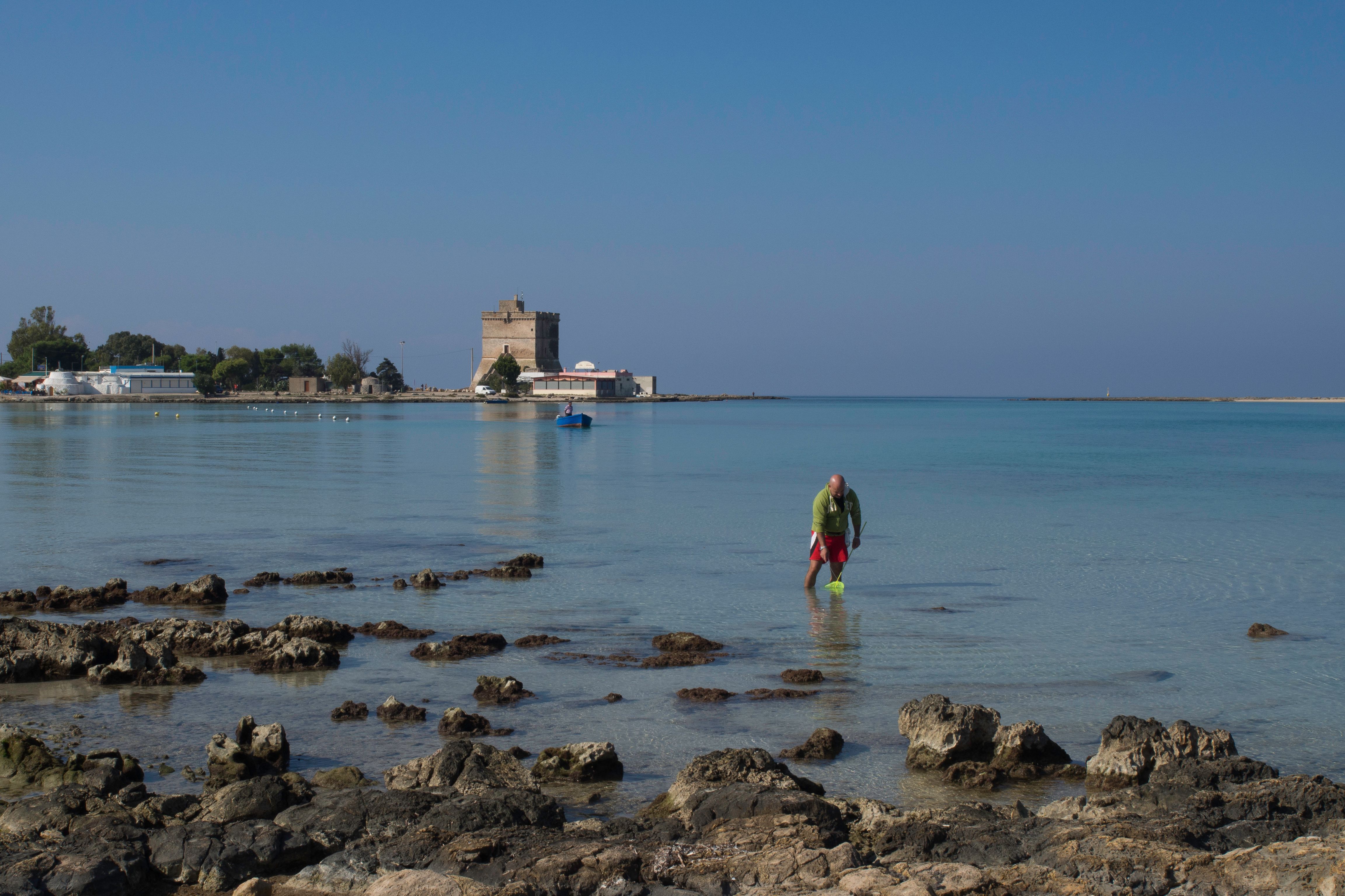 A man explores the shallow waters near the rocky shores of Sant'Isidoro, Puglia, Italy.