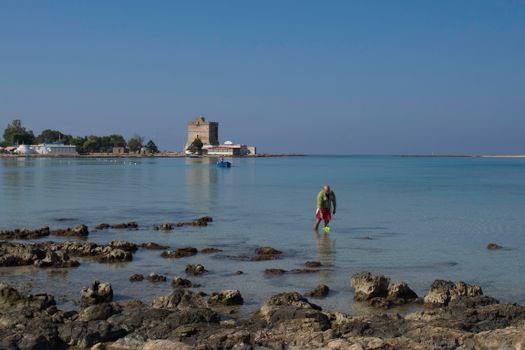 A man explores the shallow waters near the rocky shores of Sant'Isidoro, Puglia, Italy.