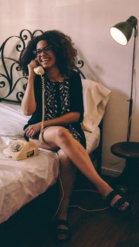 Smiling woman sitting on bed using a vintage rotary phone, emitting a relaxed and retro vibe.
