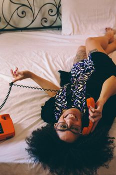 Happy woman lying on bed using a retro orange rotary phone in a cozy bedroom setting.
