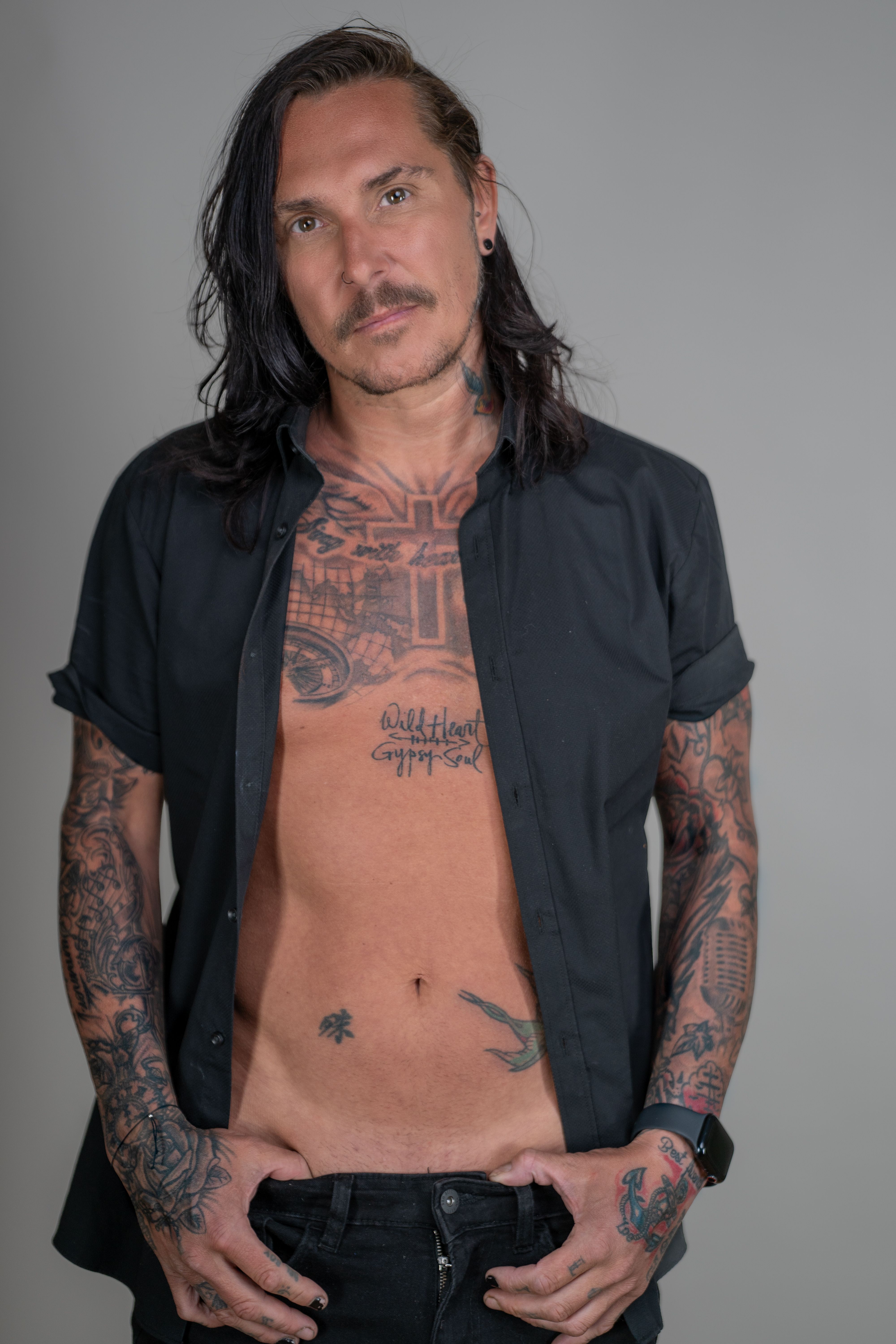 Stylish tattooed man with long hair posing confidently with an open black shirt.