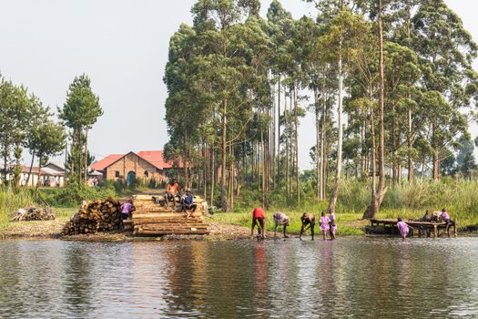 A group of people working by a lakeside with tall trees and a building in the background, showcasing rural life.