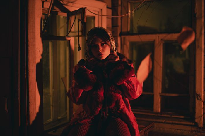 A woman in a red coat standing by a window in dim, dramatic lighting., by Daniil Kondrashin on Pexels