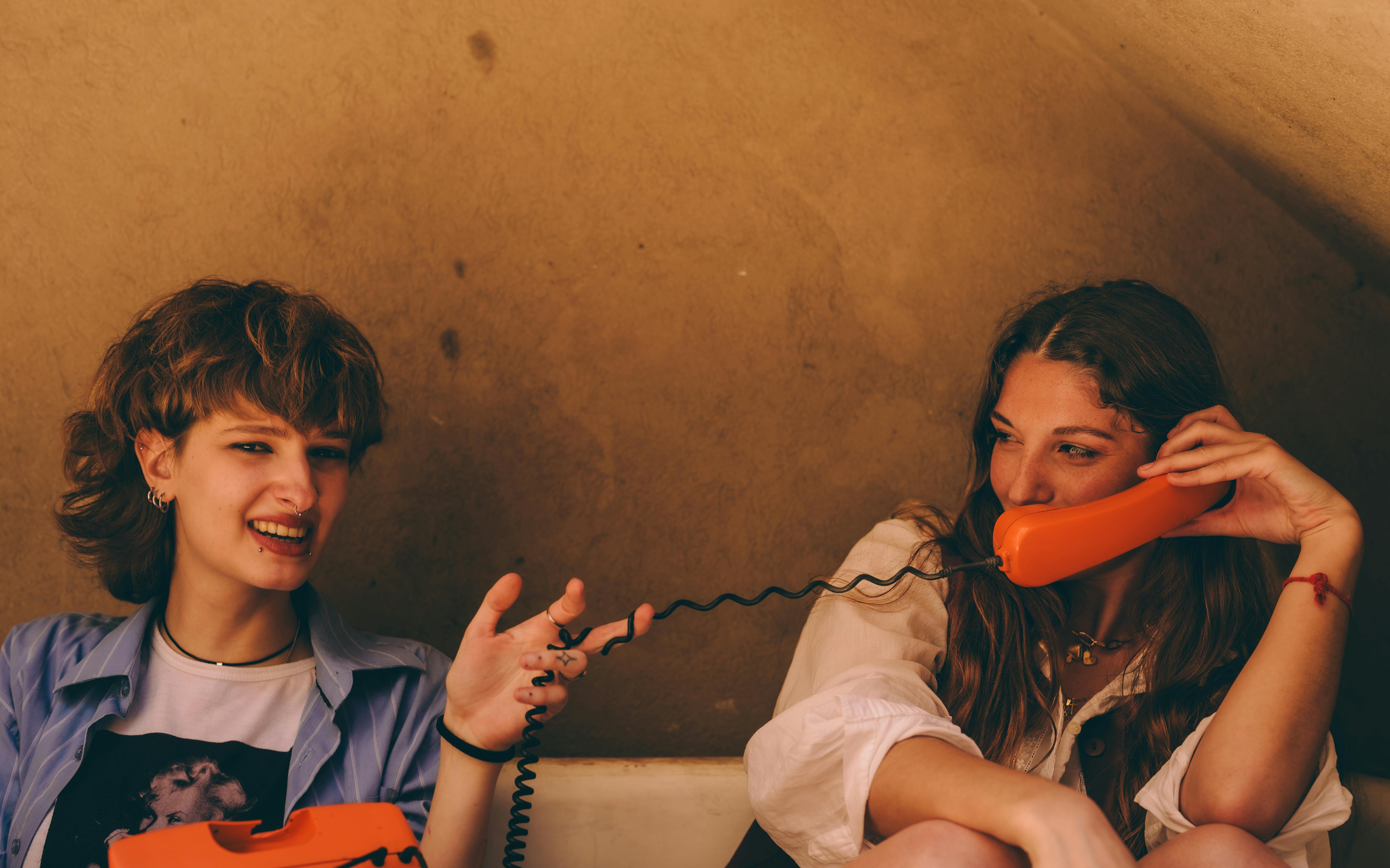 grátis Duas jovens desfrutando de uma conversa divertida usando telefones antigos laranja dentro de casa. Foto profissional
