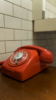 A classic red rotary telephone sits on a wooden table against a tiled wall, evoking nostalgia.