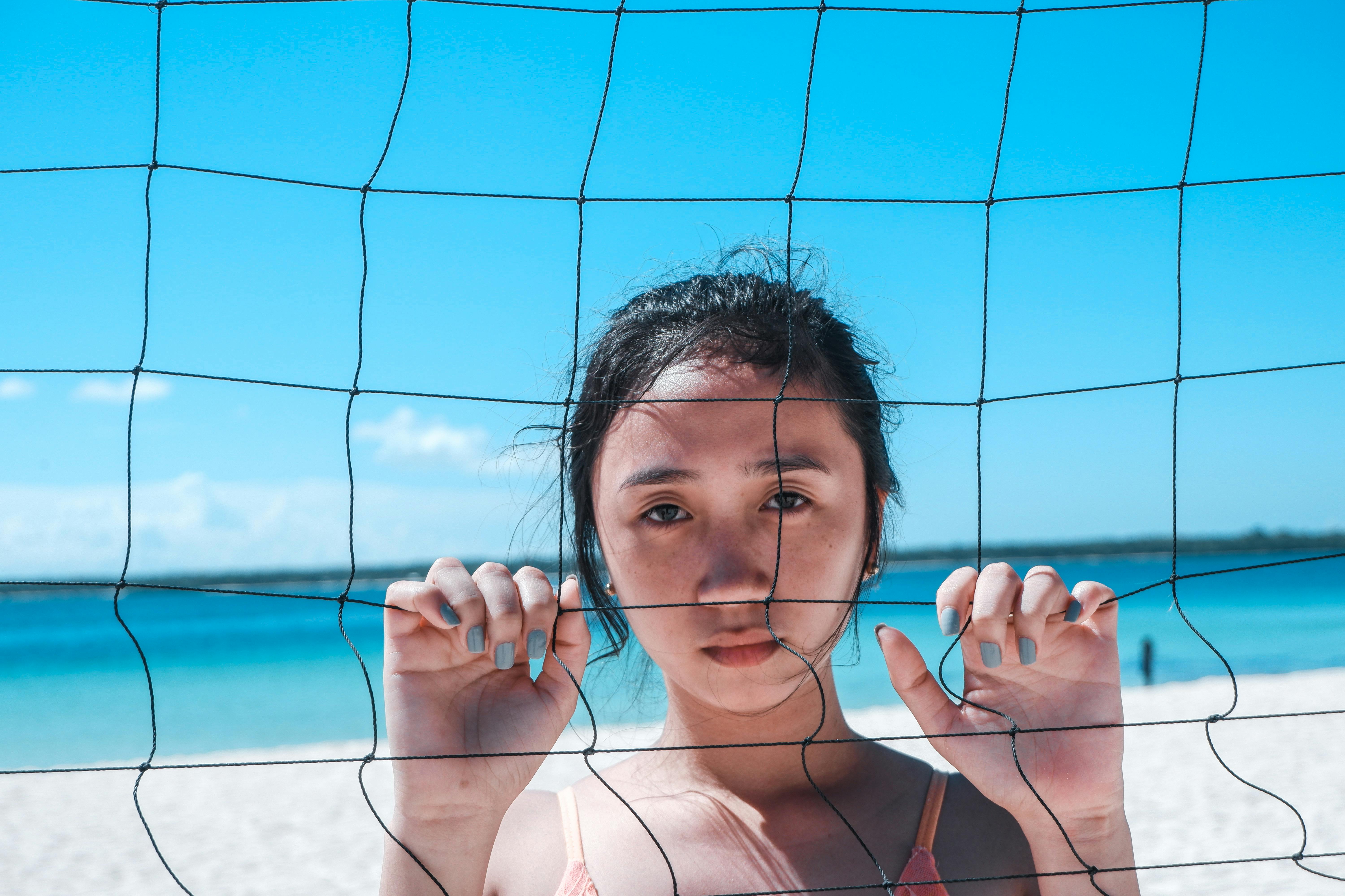 一个阳光明媚的日子，海边沙滩排球网后站着一位若有所思的年轻女子