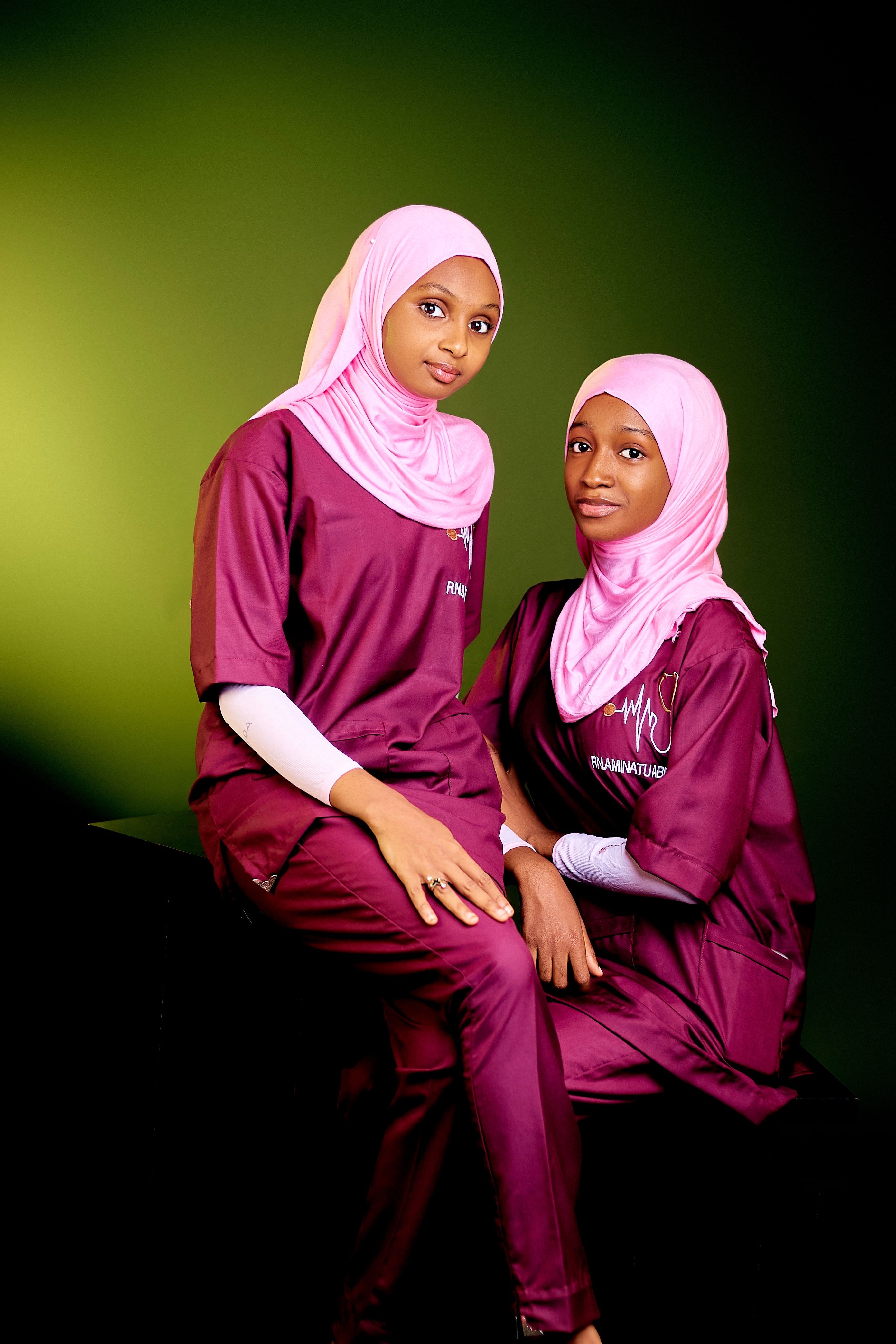 Two women wearing hijabs and scrubs posing against a green backdrop.