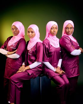 Four women in medical uniforms and pink hijabs pose confidently against a studio backdrop.