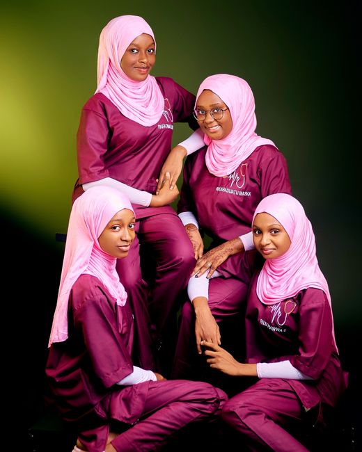 Four women in matching hijabs and burgundy outfits pose with a friendly demeanor.