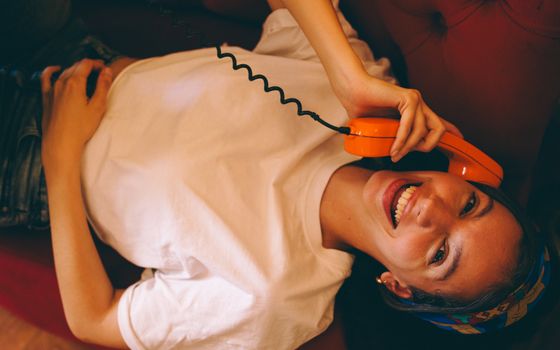 Happy woman lying down, chatting on a retro orange phone, enjoying a relaxed conversation indoors.