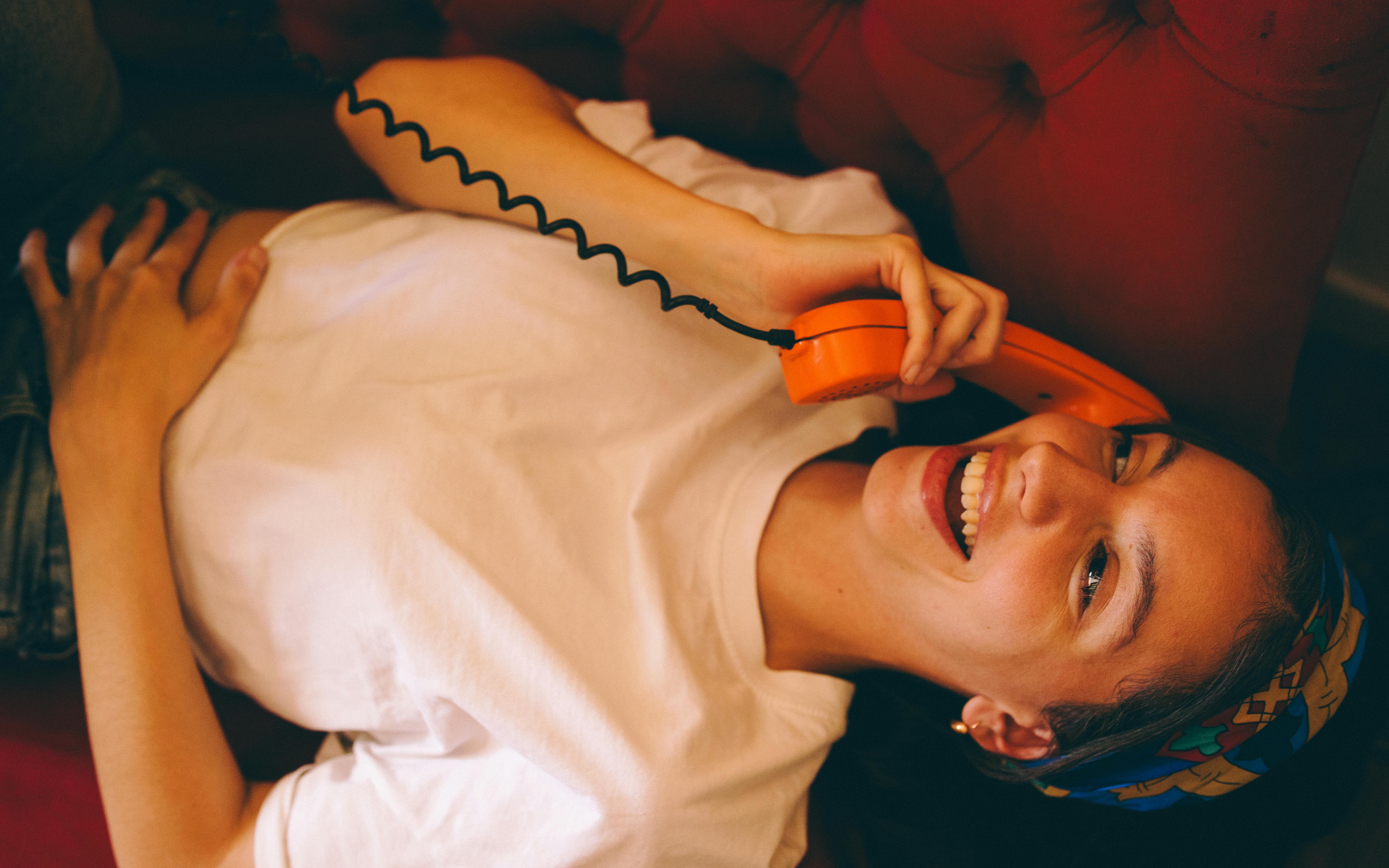 Gratuit O femeie zâmbitoare, întinsă pe o canapea, ține în mână un telefon portocaliu vintage, în interior. Fotografie de stoc