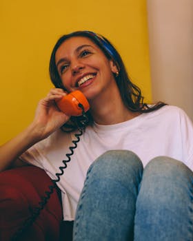 Smiling woman talks on a vintage orange telephone indoors with a cheerful expression.