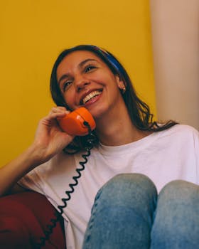 Smiling young woman talking on a classic orange rotary phone indoors.