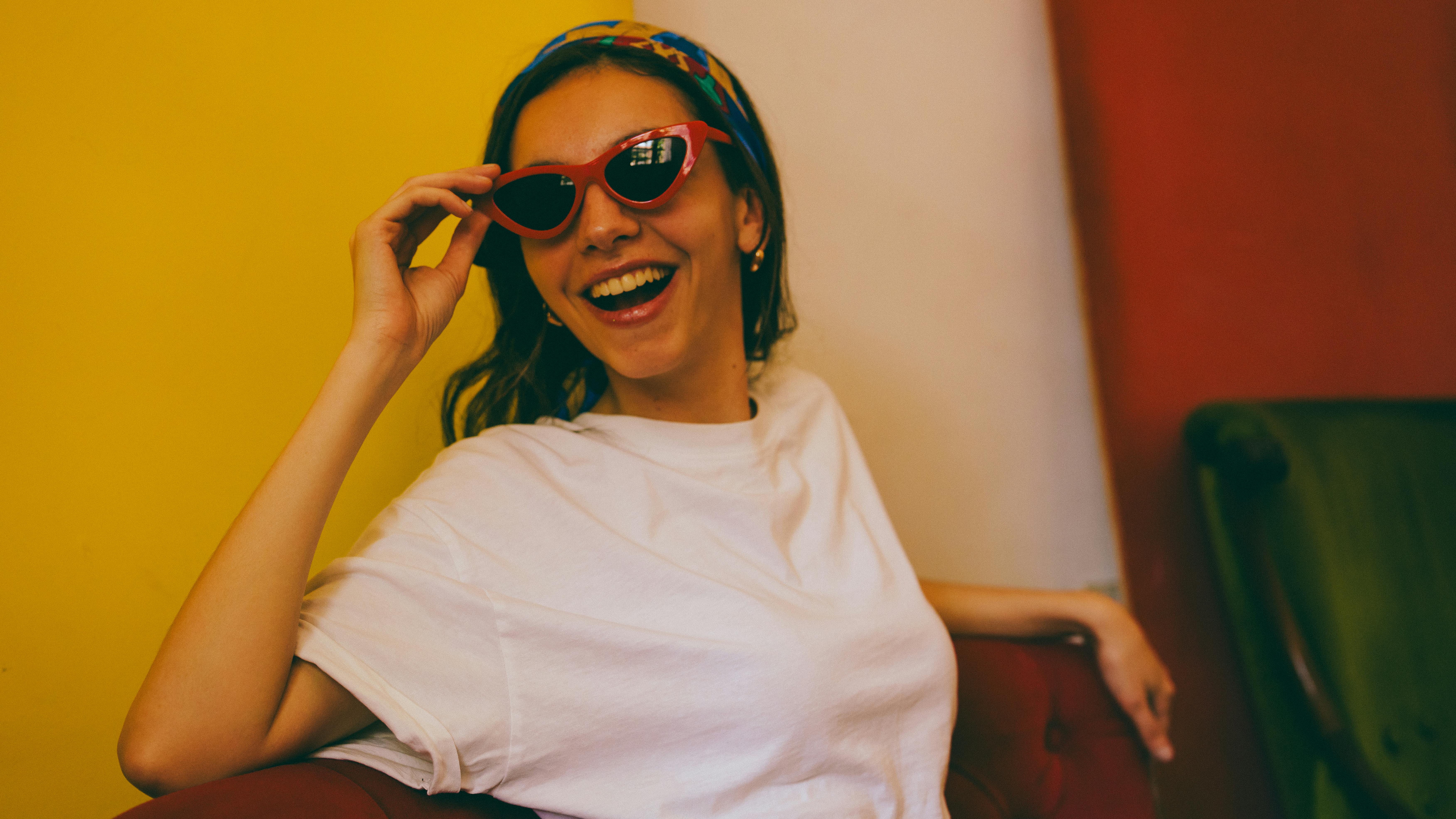 grátis Mulher alegre usando óculos de sol vermelhos em frente a uma parede colorida, irradiando felicidade. Foto profissional