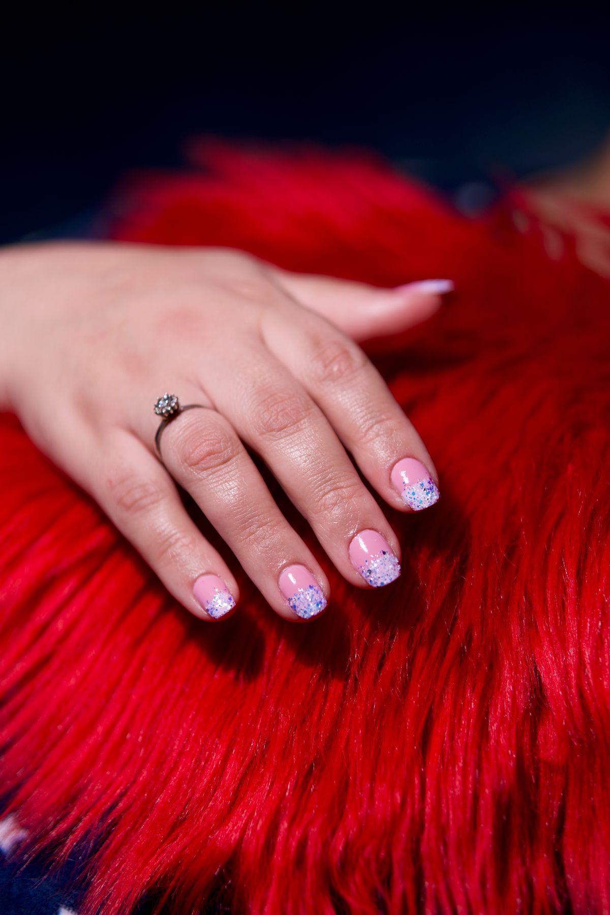 A woman's hand with a glittery pink manicure rests on vibrant red fur, showcasing a stylish ring.