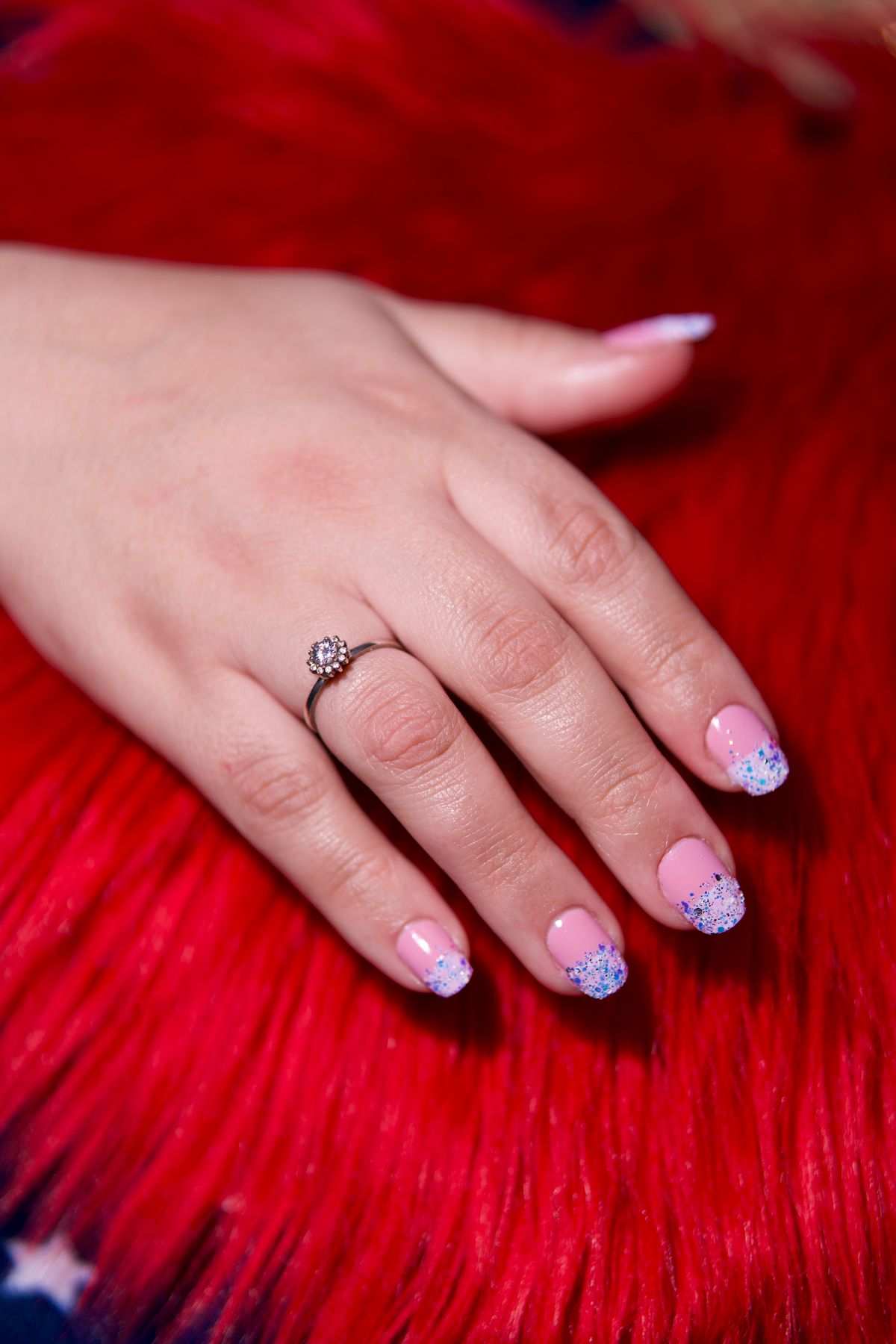Close-up of a woman's hand with a ring and glittery nail polish on a vibrant red background.