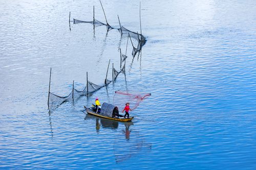 Free Two fishermen on a small boat casting fishing nets on a tranquil blue water surface. Stock Photo