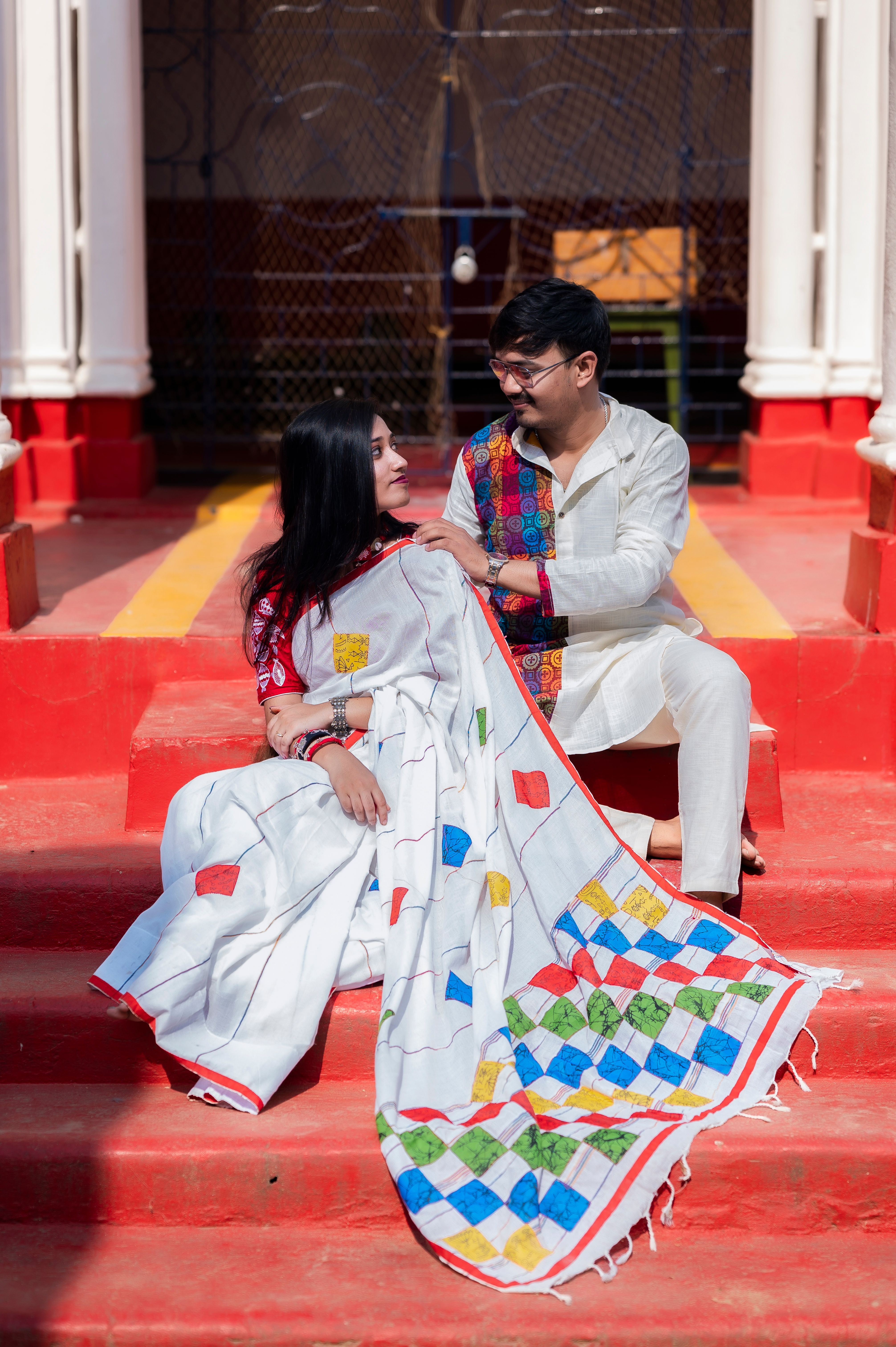 A couple in vibrant Indian attire sitting on red steps, exuding cultural charm and elegance.