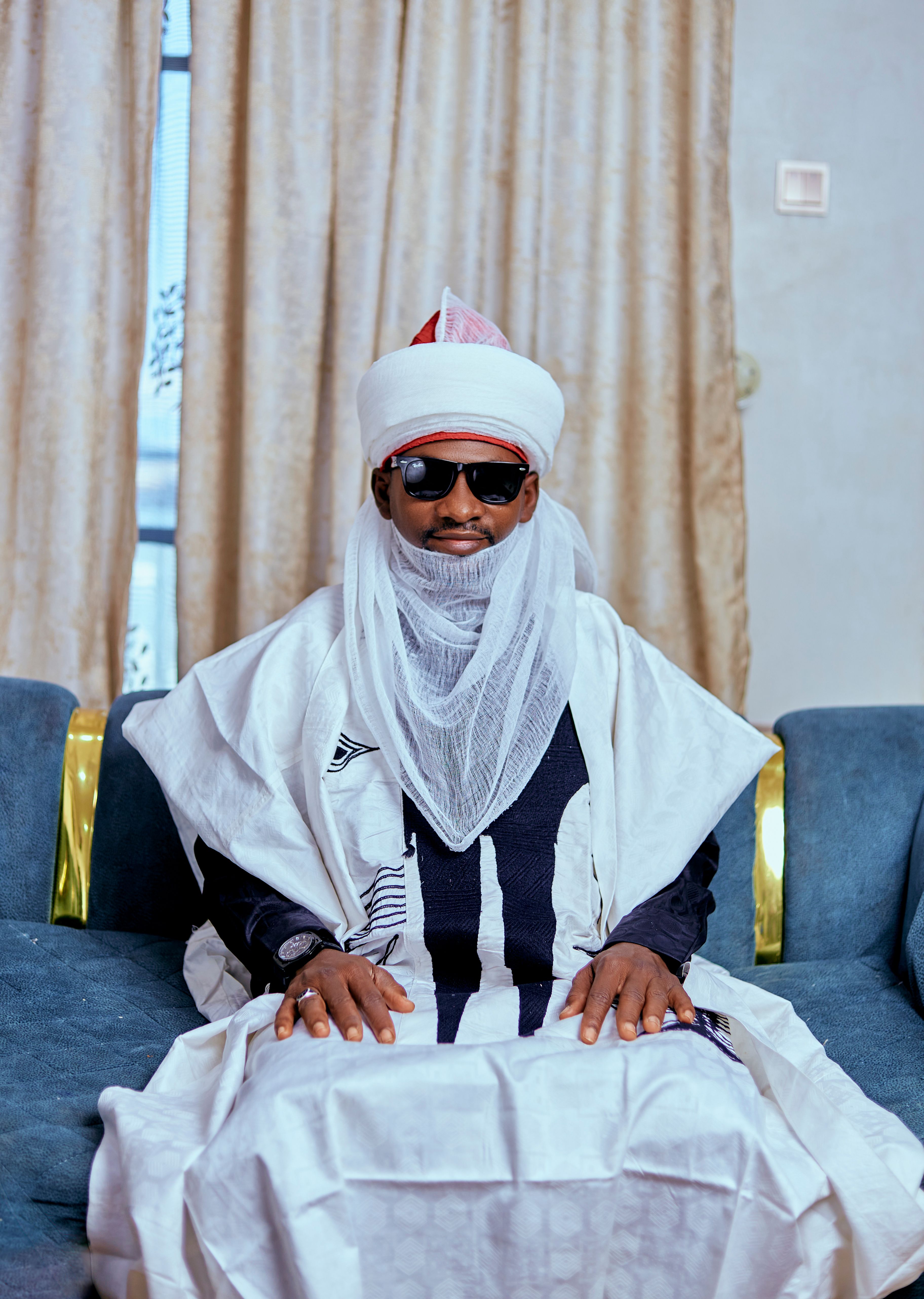 A man in traditional attire with sunglasses sits indoors against a neutral background.