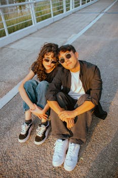 Casual young couple wearing sunglasses sitting on a bridge, enjoying the warm sunset outdoors.
