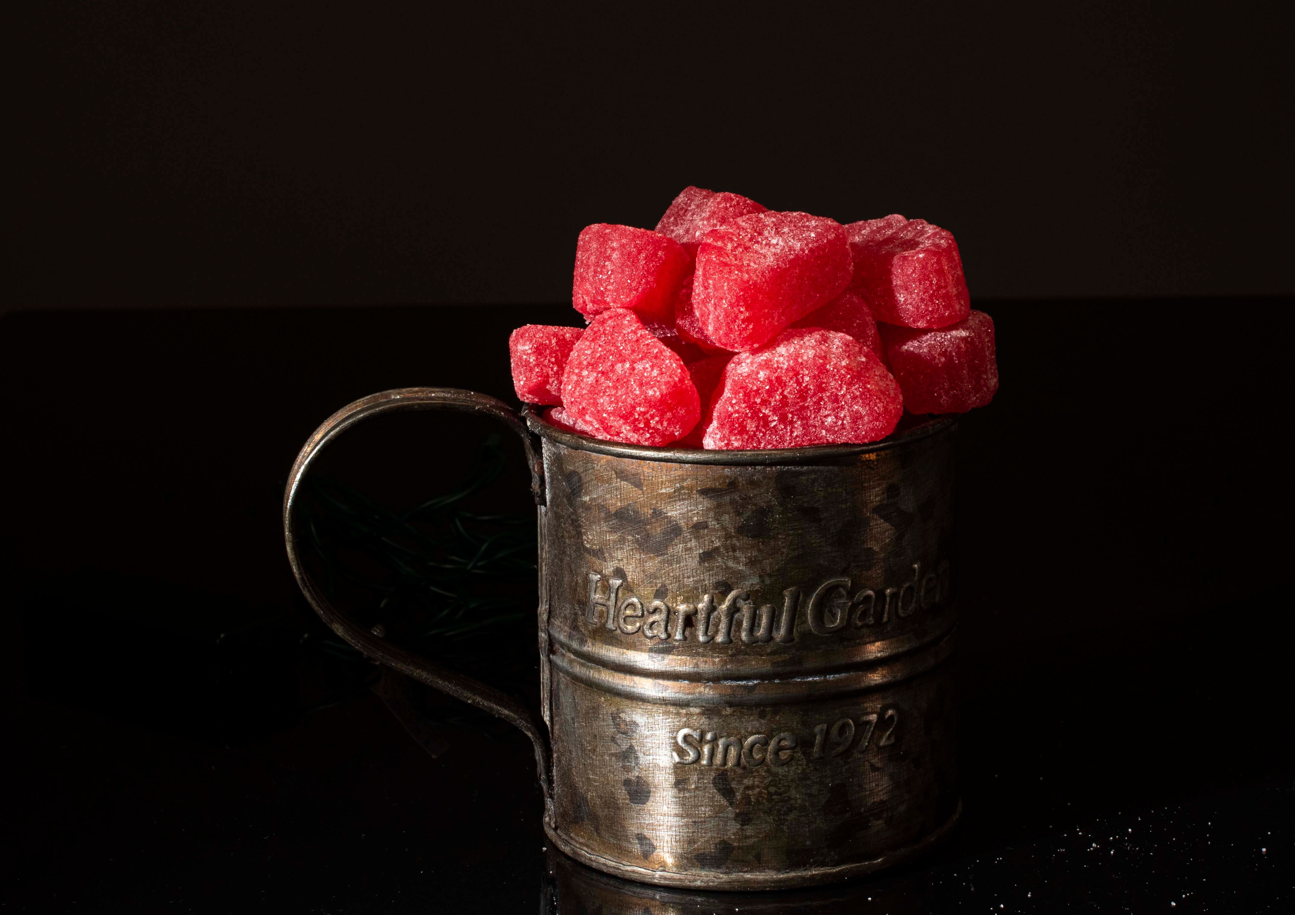 A rustic metal cup filled with vibrant red sugar-coated candies on a dark background.