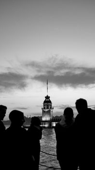 Silhouetted figures gaze at the illuminated Maiden's Tower surrounded by calm waters at twilight.