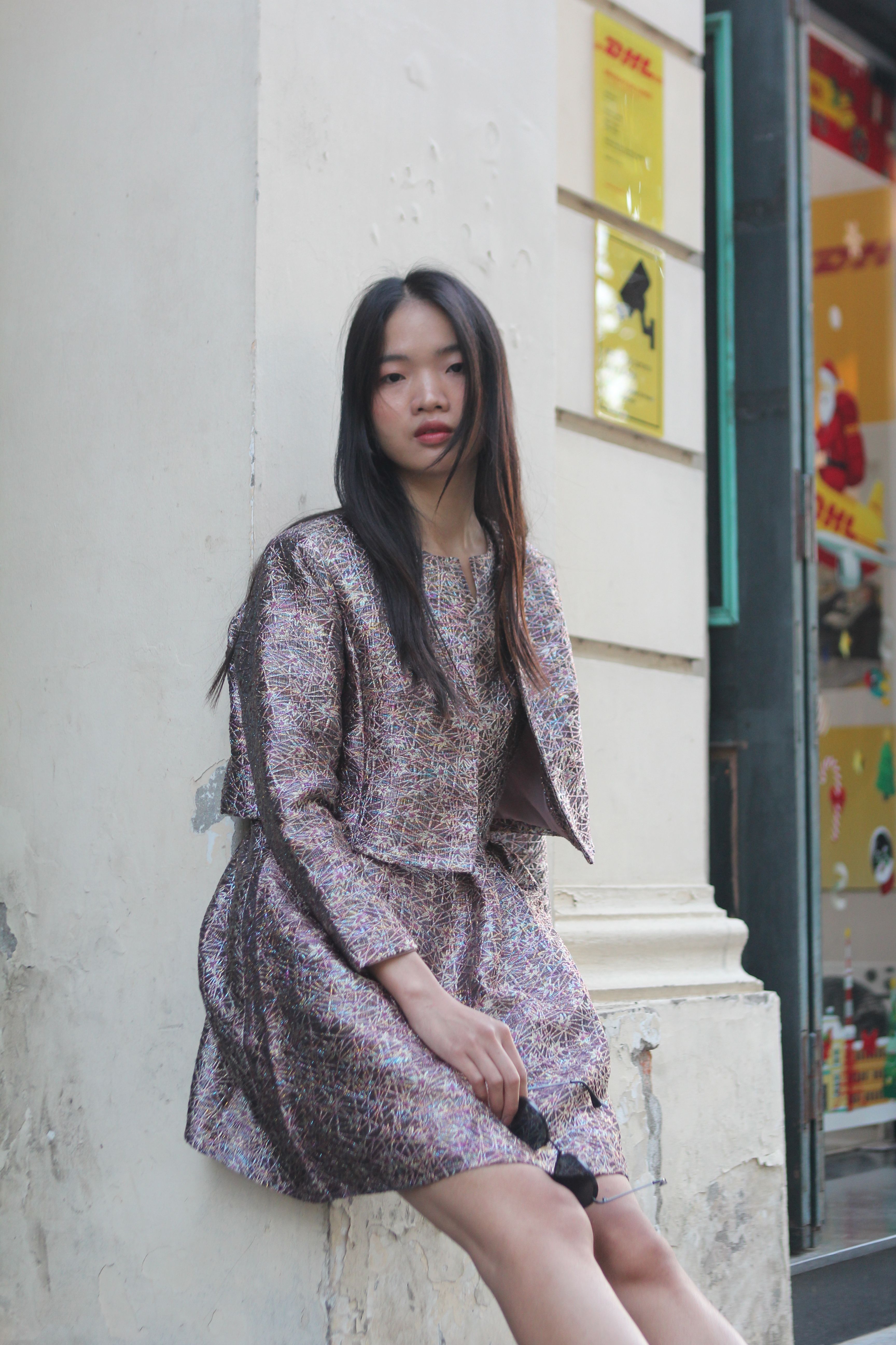 A fashionable young woman poses outdoors in a stylish outfit, leaning against a wall.