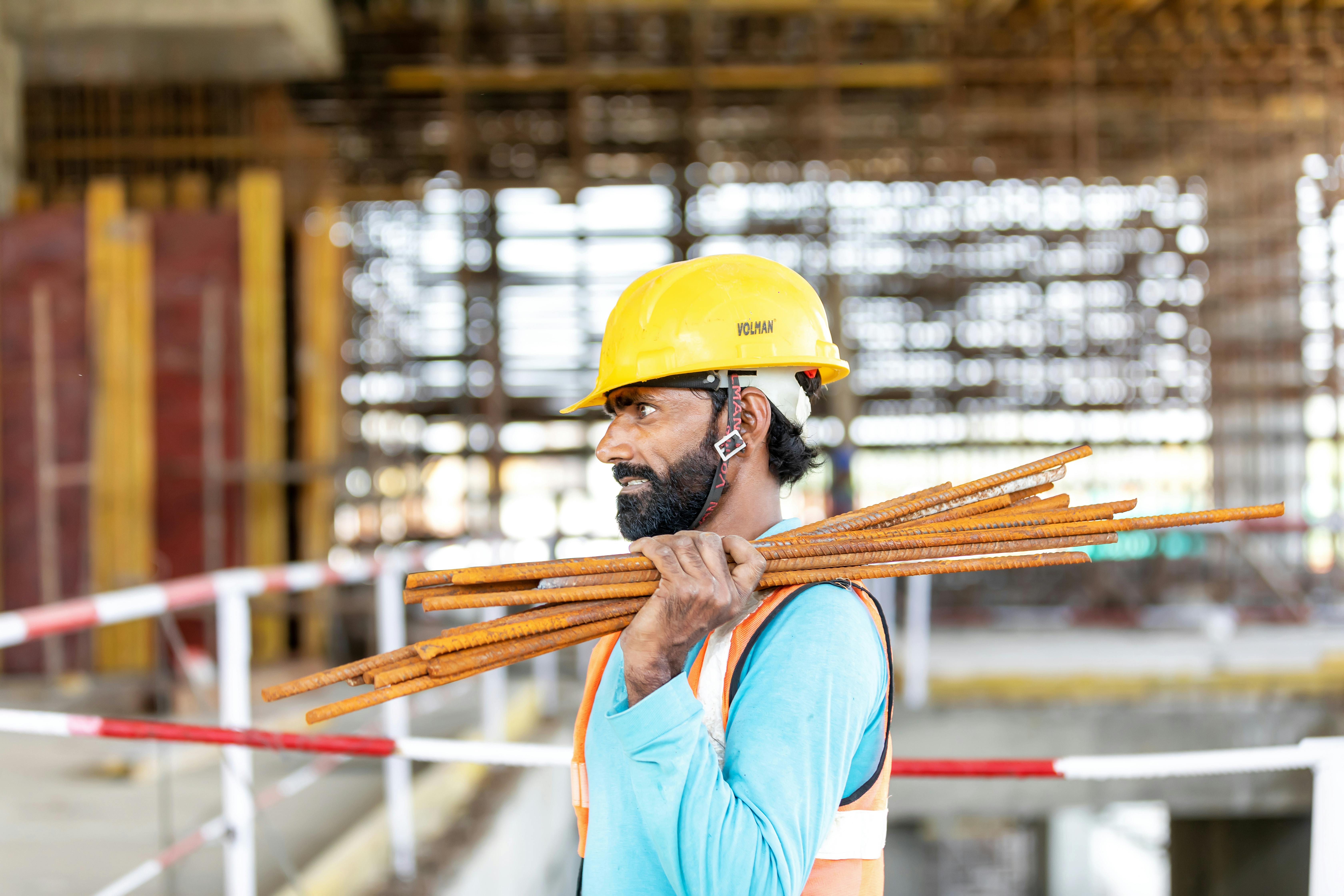 Construction worker at a building site carrying rebar, wearing a safety helmet and vest.