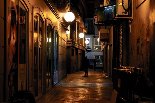 A dark, narrow urban alleyway illuminated by warm streetlights at night, creating a moody atmosphere.