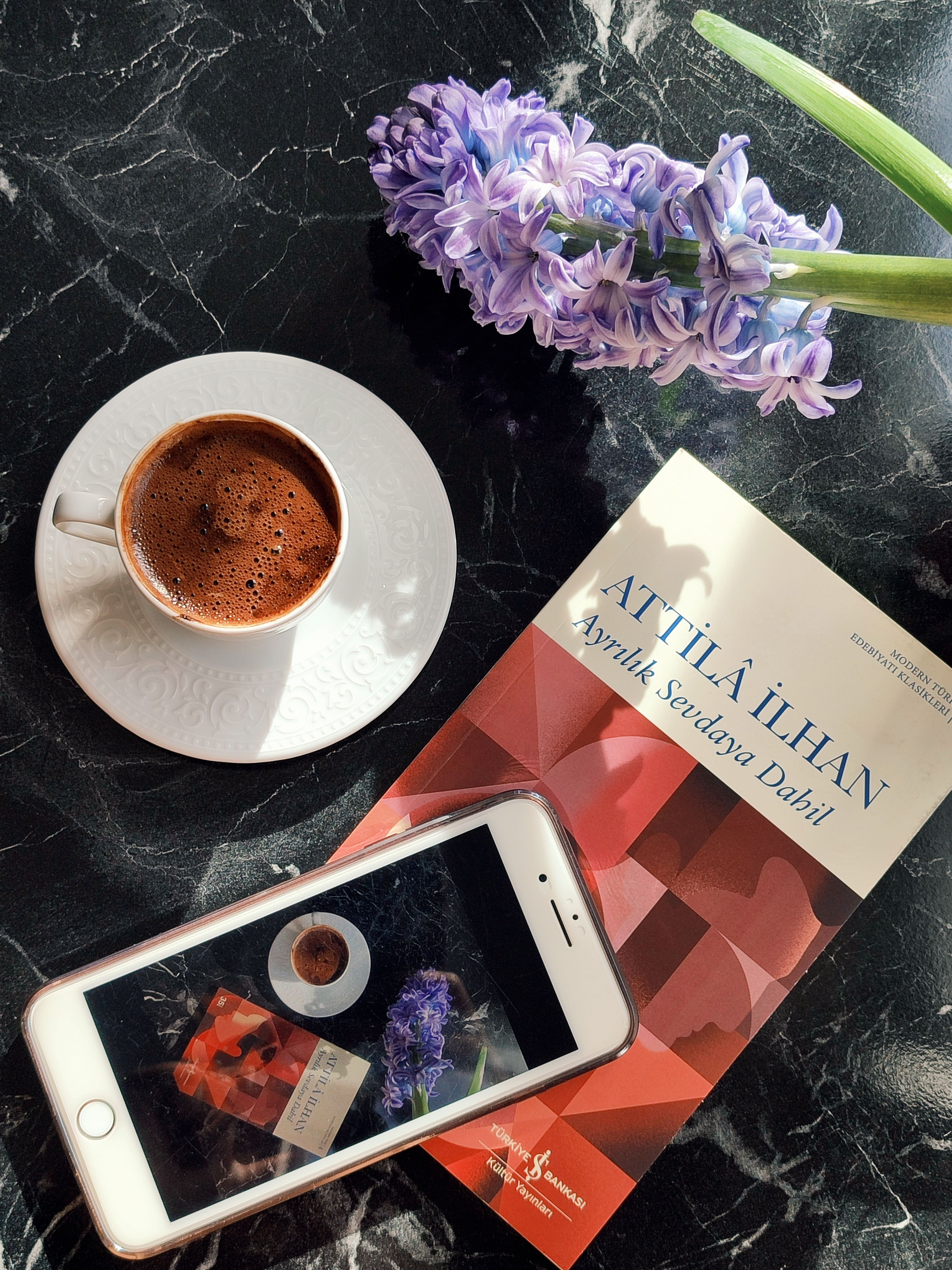 A cozy morning scene with coffee, a book, flowers, and a smartphone on a marble surface.