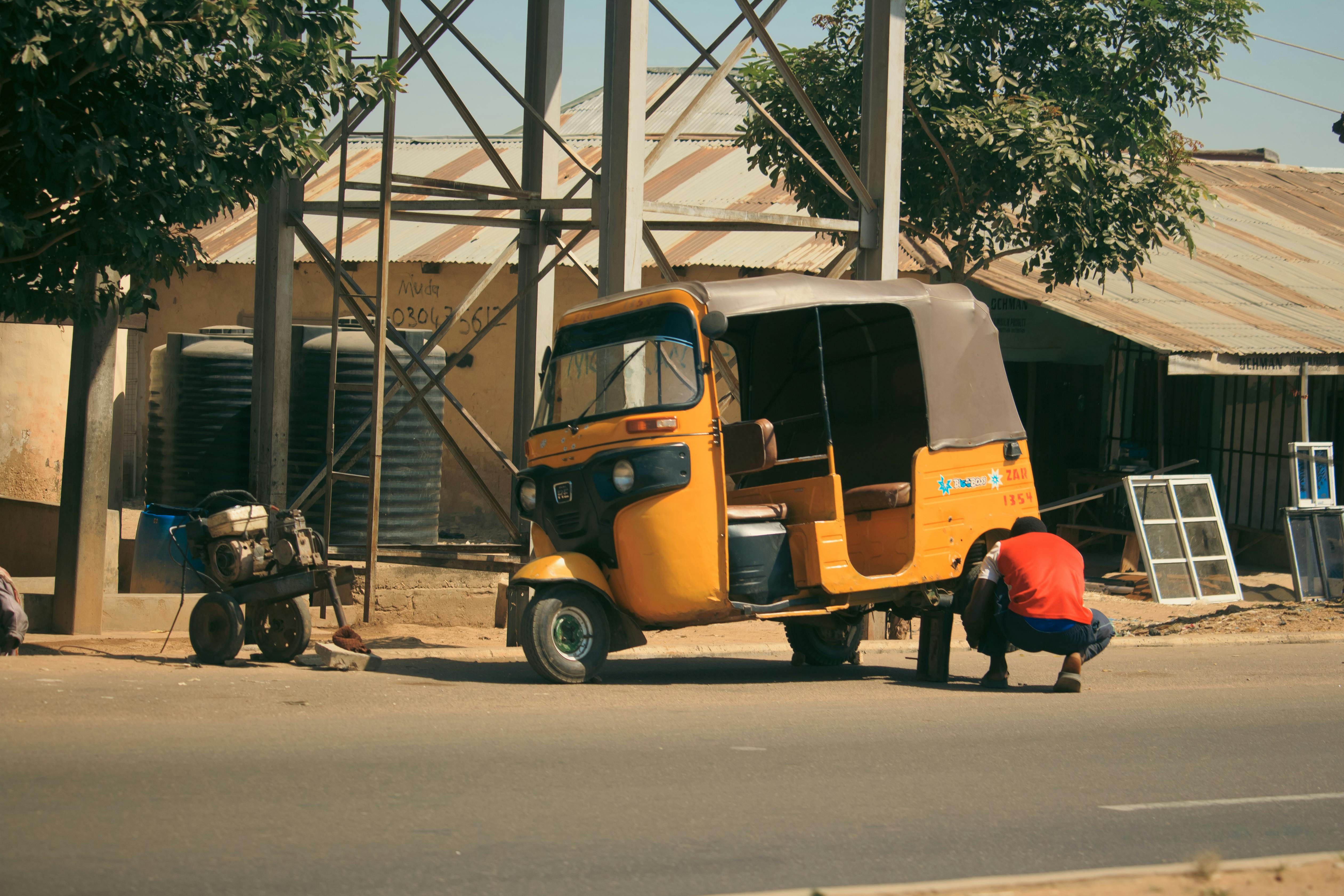 A mechanic repairs a yellow tuk-tuk on a city street under a sunny sky. Urban scene.