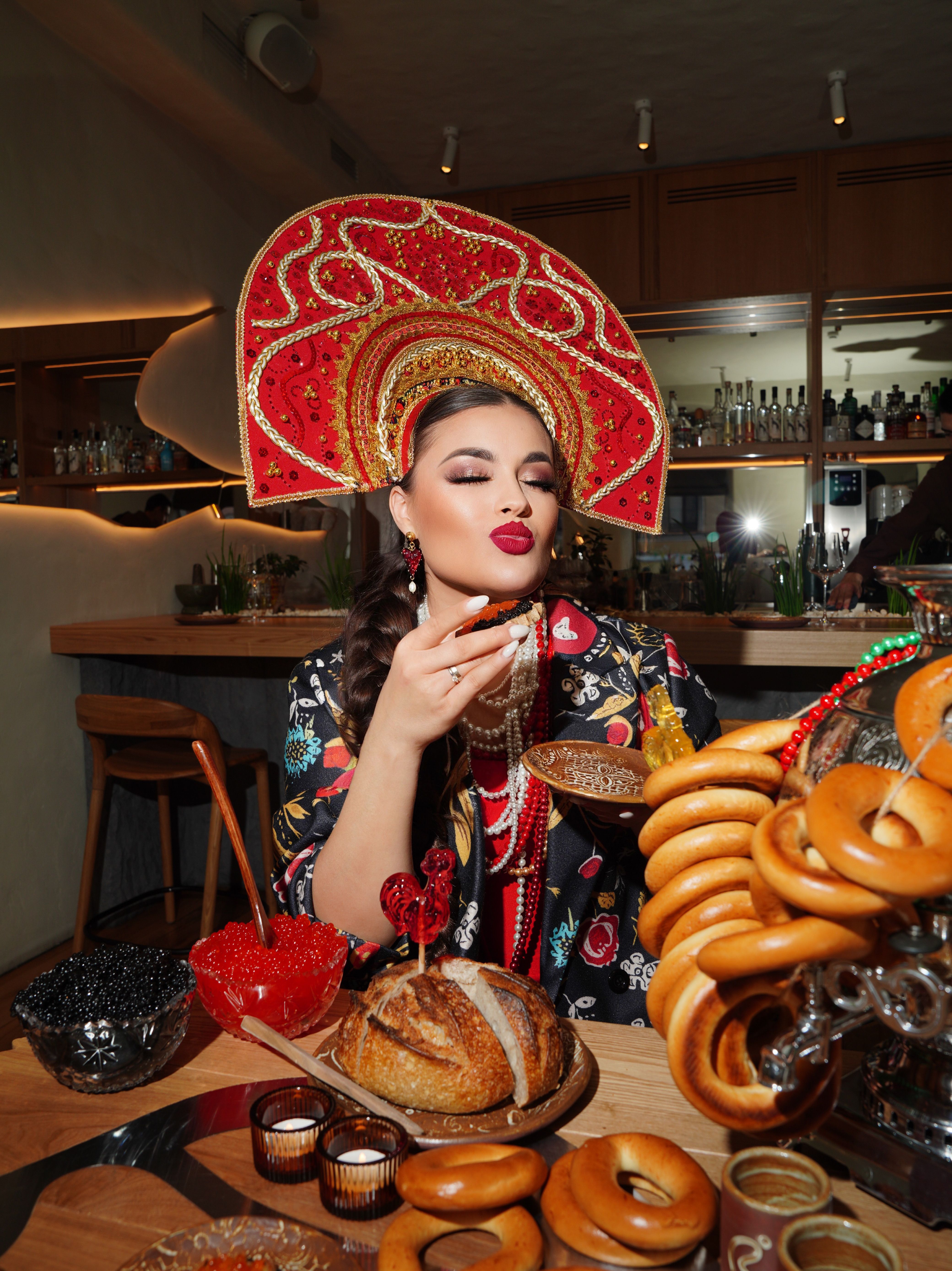 A woman wearing a decorative headdress enjoying traditional Russian pastries indoors.