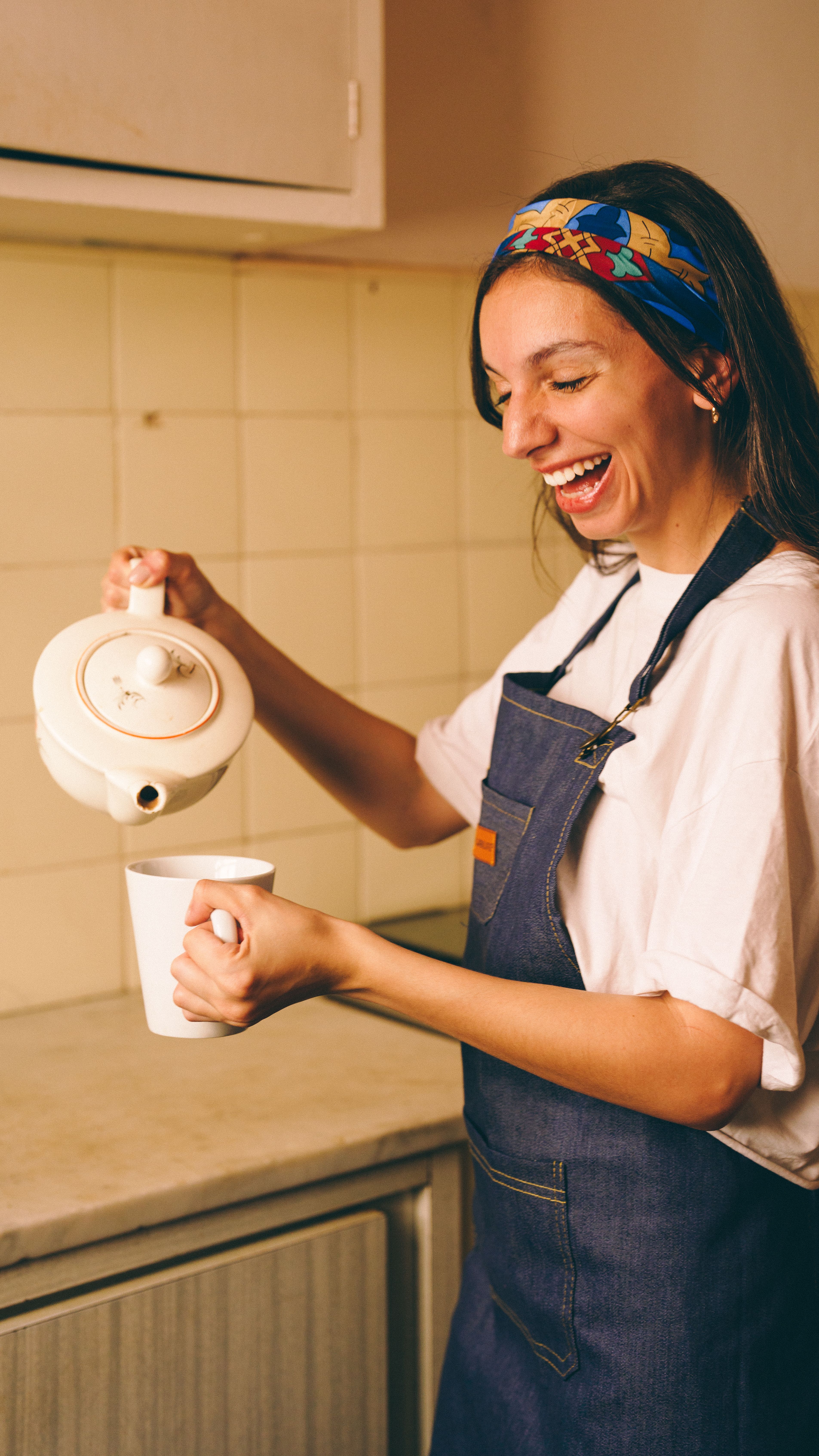 A cheerful woman in a kitchen pouring tea from a teapot into a cup, wearing an apron and colorful headband.