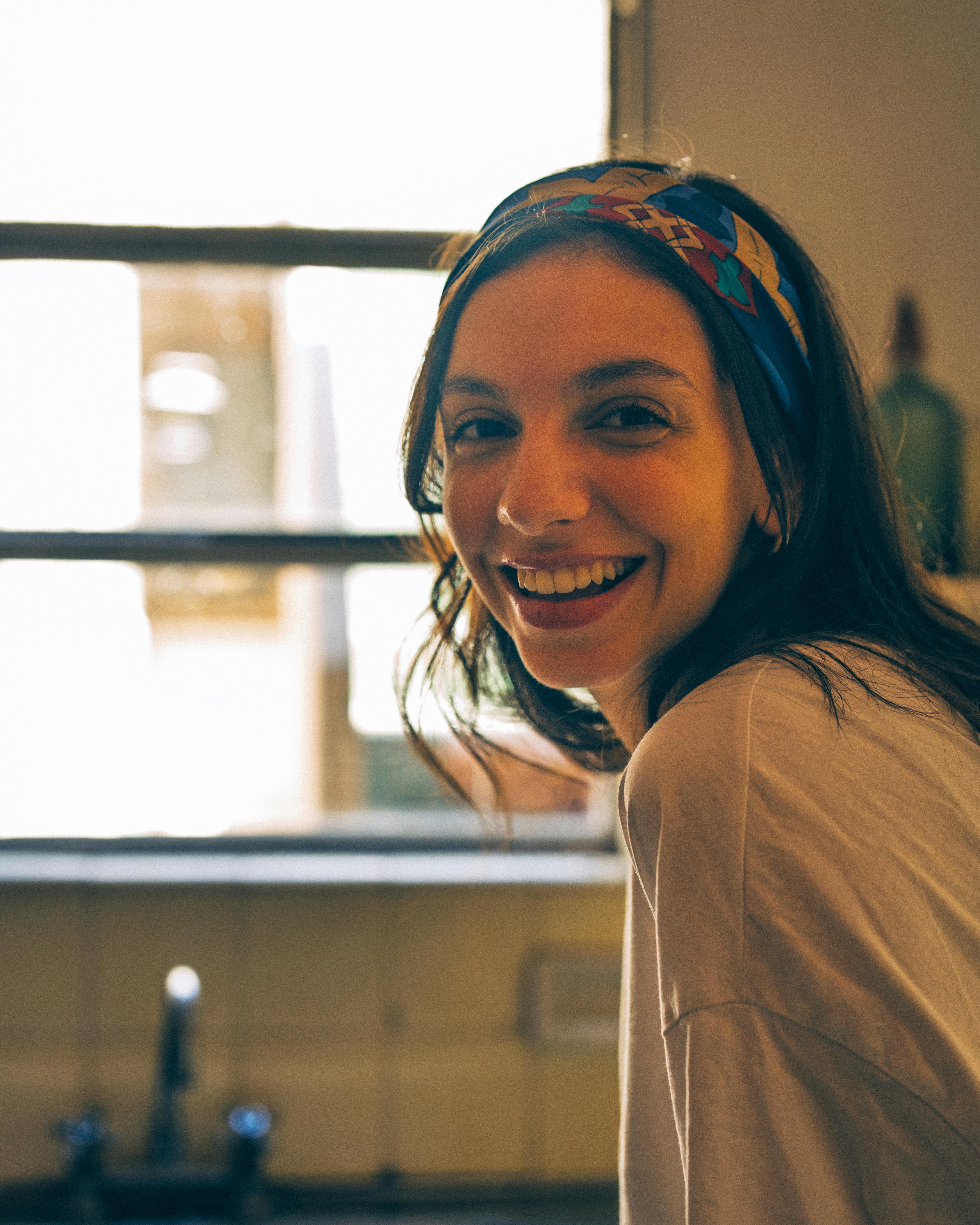 무료 한 여성이 햇살이 비치는 주방에 앉아 다채로운 머리띠를 두르고 따뜻하게 미소 짓고 있습니다. 스톡 사진