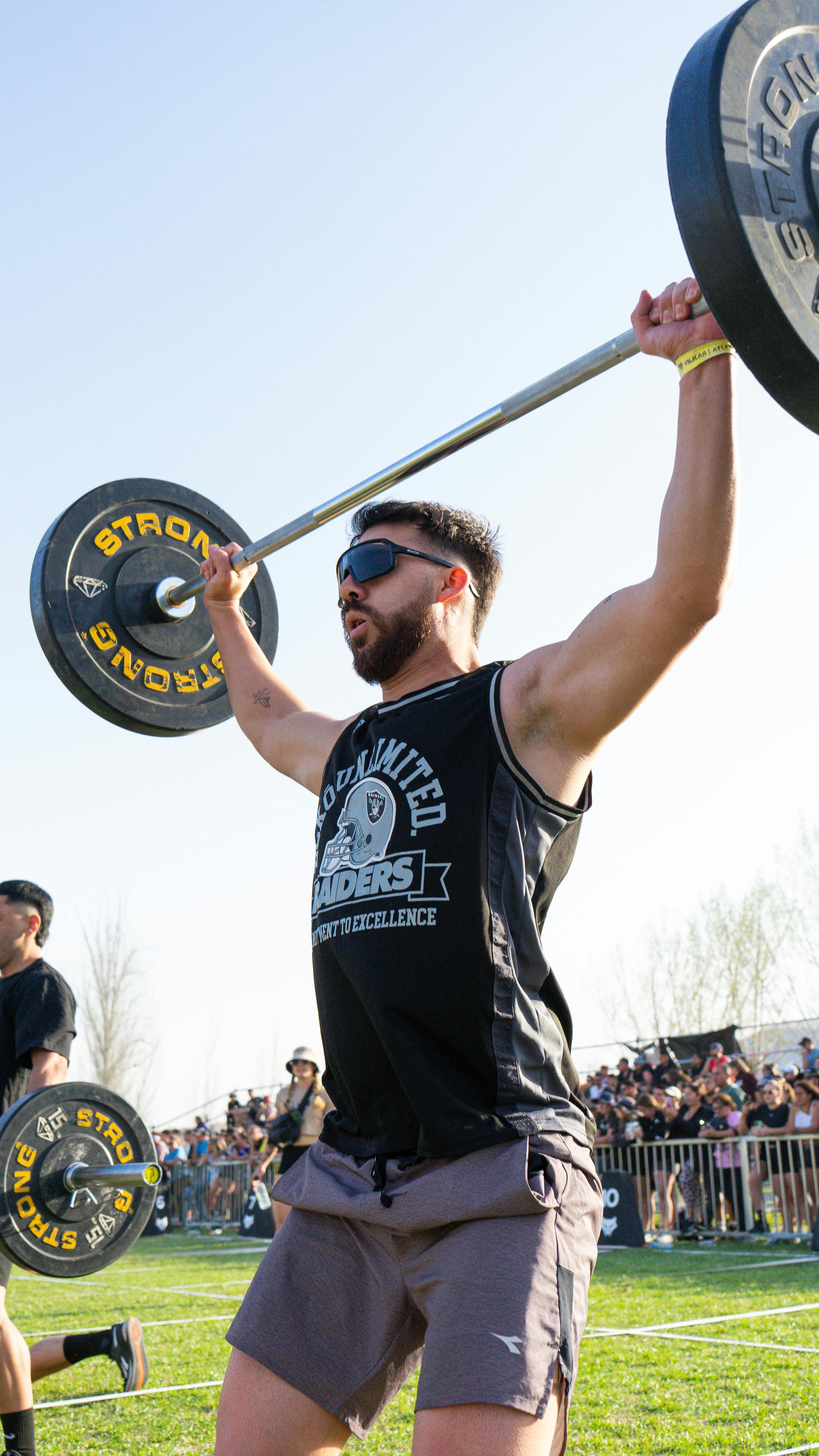 A man participating in a fitness competition lifting a barbell outdoors on a sunny day.