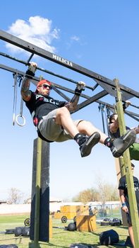 Athletes perform pull-ups on an outdoor obstacle course under blue skies.