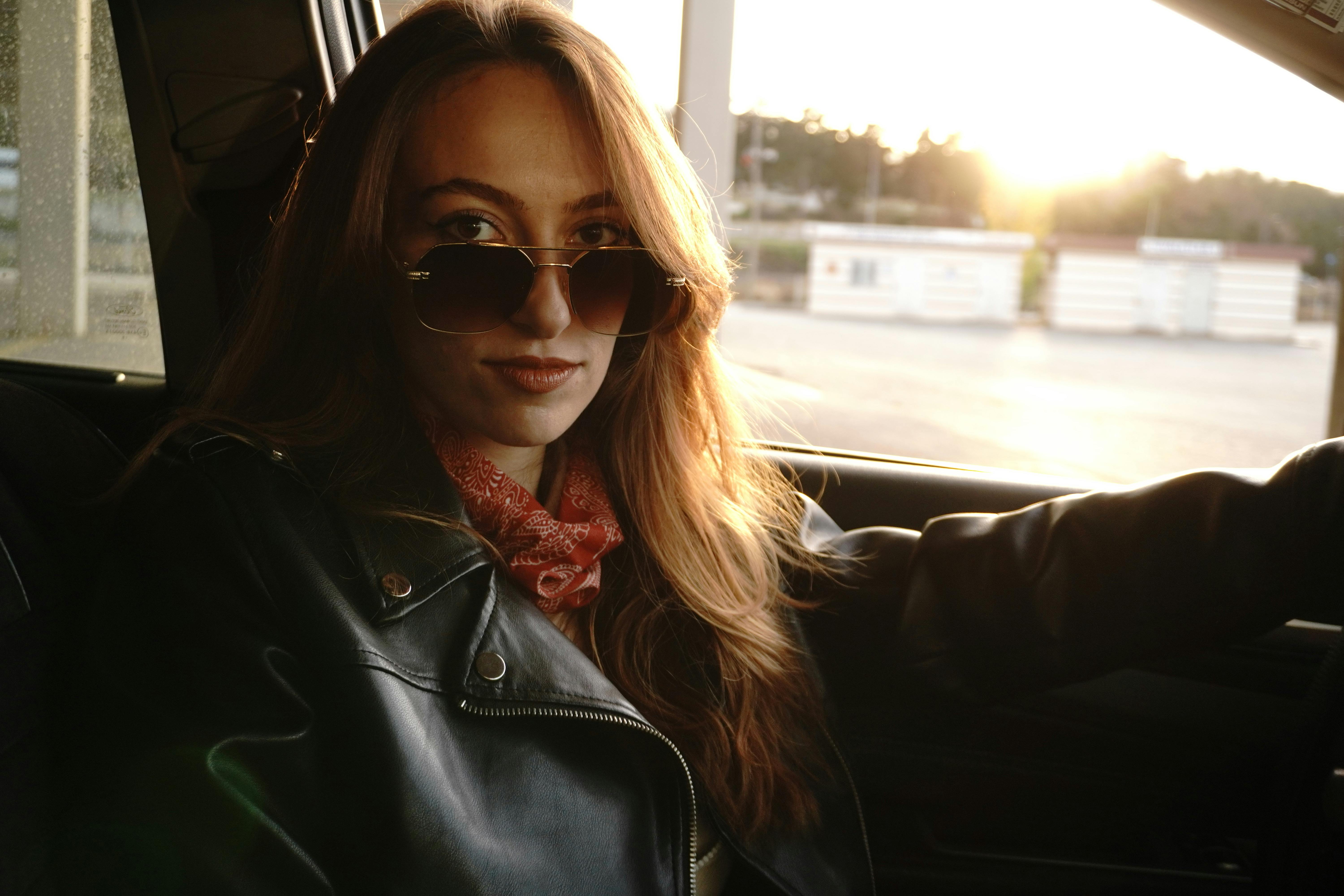 Stylish woman in a car during sunset wearing sunglasses and leather jacket.