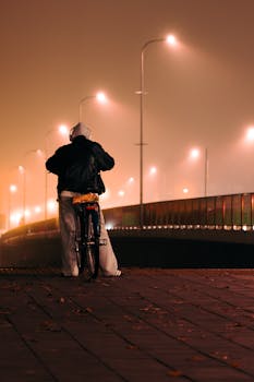 Silhouette of a cyclist on a foggy night bridge, with glowing streetlights casting a moody atmosphere.