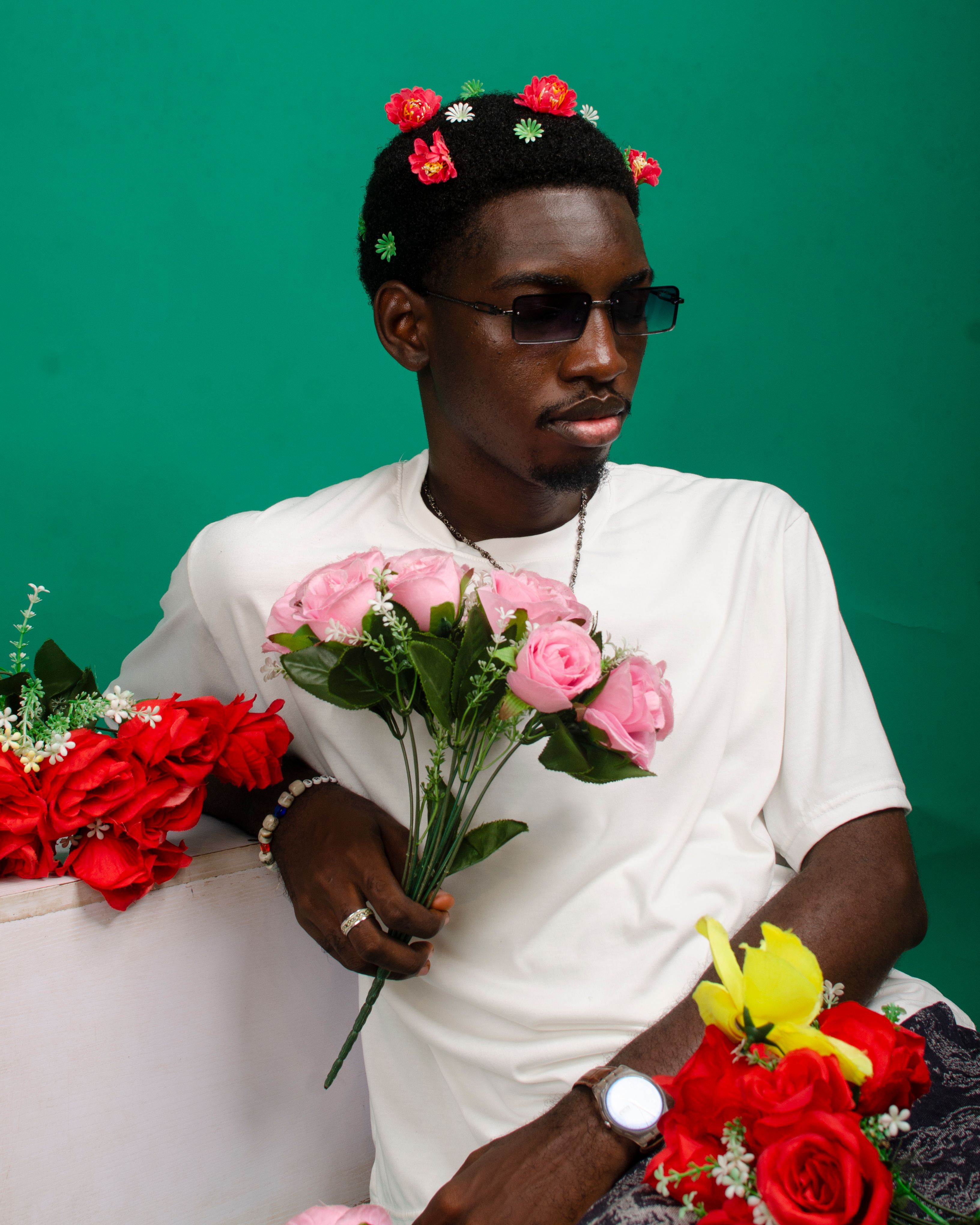 Fashionable man in sunglasses poses with colorful flowers, exuding confidence against a green backdrop.