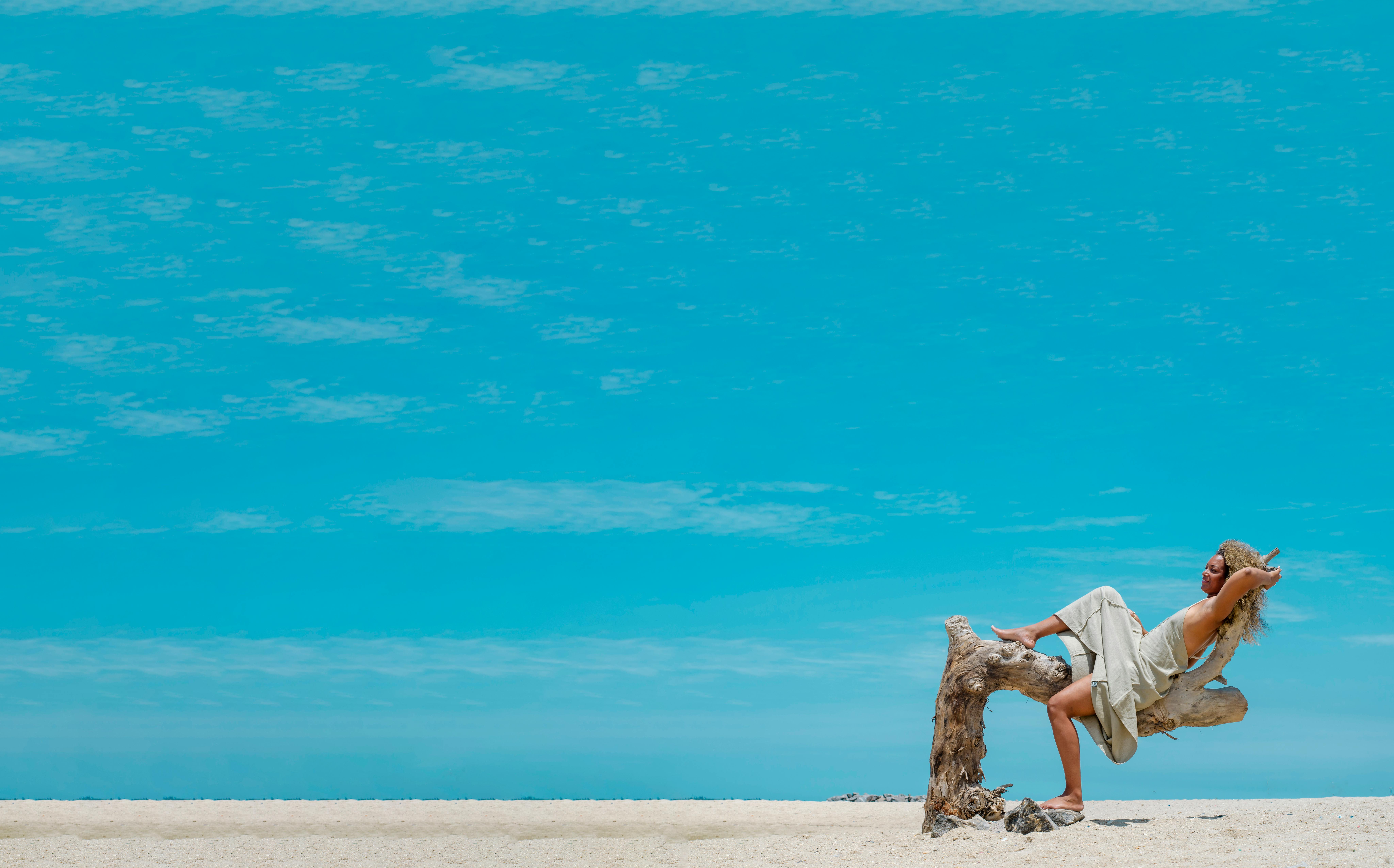 A serene scene of a woman lounging on driftwood on a sandy beach under a clear blue sky.