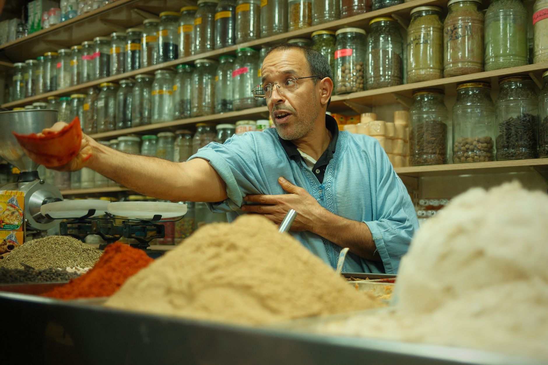 Photo : Thomas balabaud / Pexels A Moroccan man selling spices in a colorful traditional market stall, showcasing various herbs and spices.