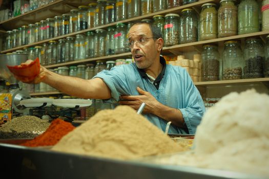 A Moroccan man selling spices in a colorful traditional market stall, showcasing various herbs and spices.