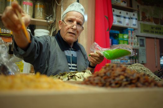 Elderly vendor at a Moroccan spice market offers vibrant spices and herbs, showcasing local culture.