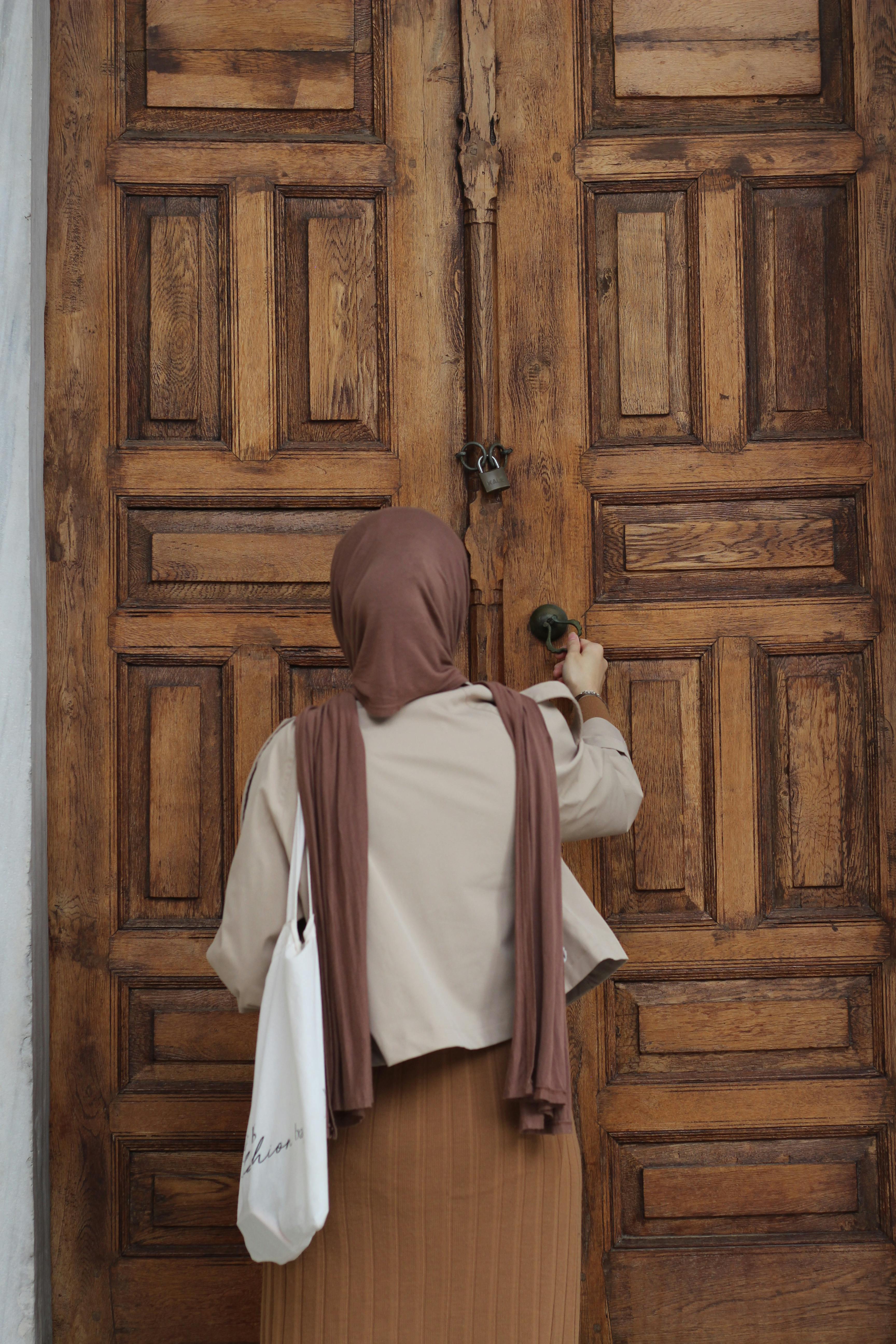 Rear view of a woman in hijab knocking on a large wooden door, symbolizing mystery or invitation.