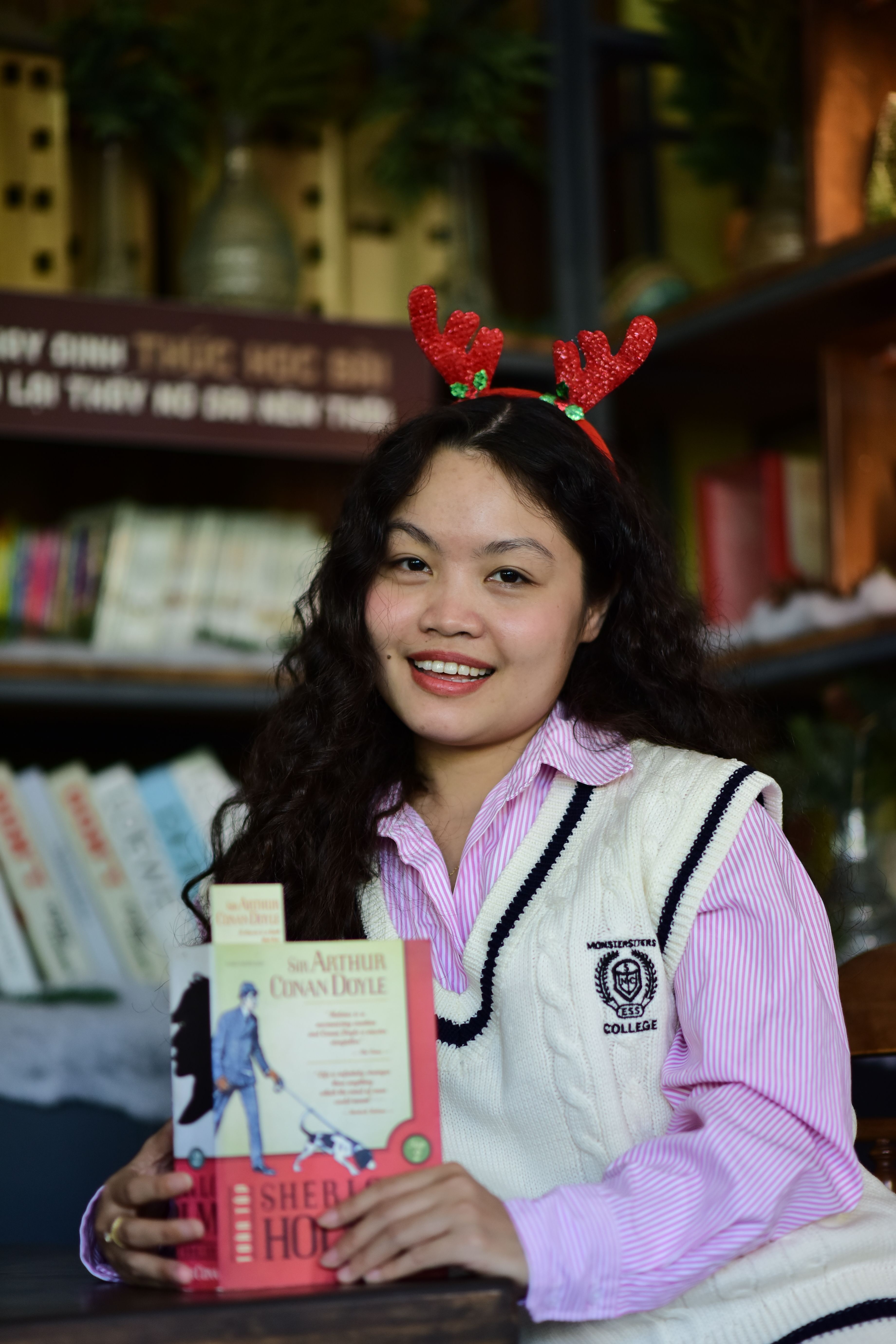 Woman indoors wearing red antler headband, holding Sherlock Holmes book, smiling.