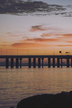 Explore a tranquil sunset with a panoramic view of a bridge and distant cityscape over calm waters.