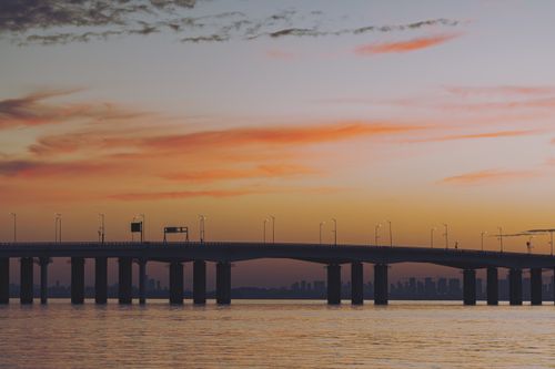 Free A breathtaking sunset illuminates a serene bridge spanning calm waters with a distant city skyline. Stock Photo