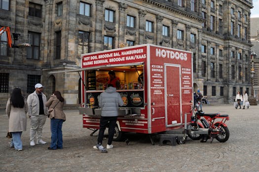 A bustling street food stall selling hot dogs in a European square with people gathered around.