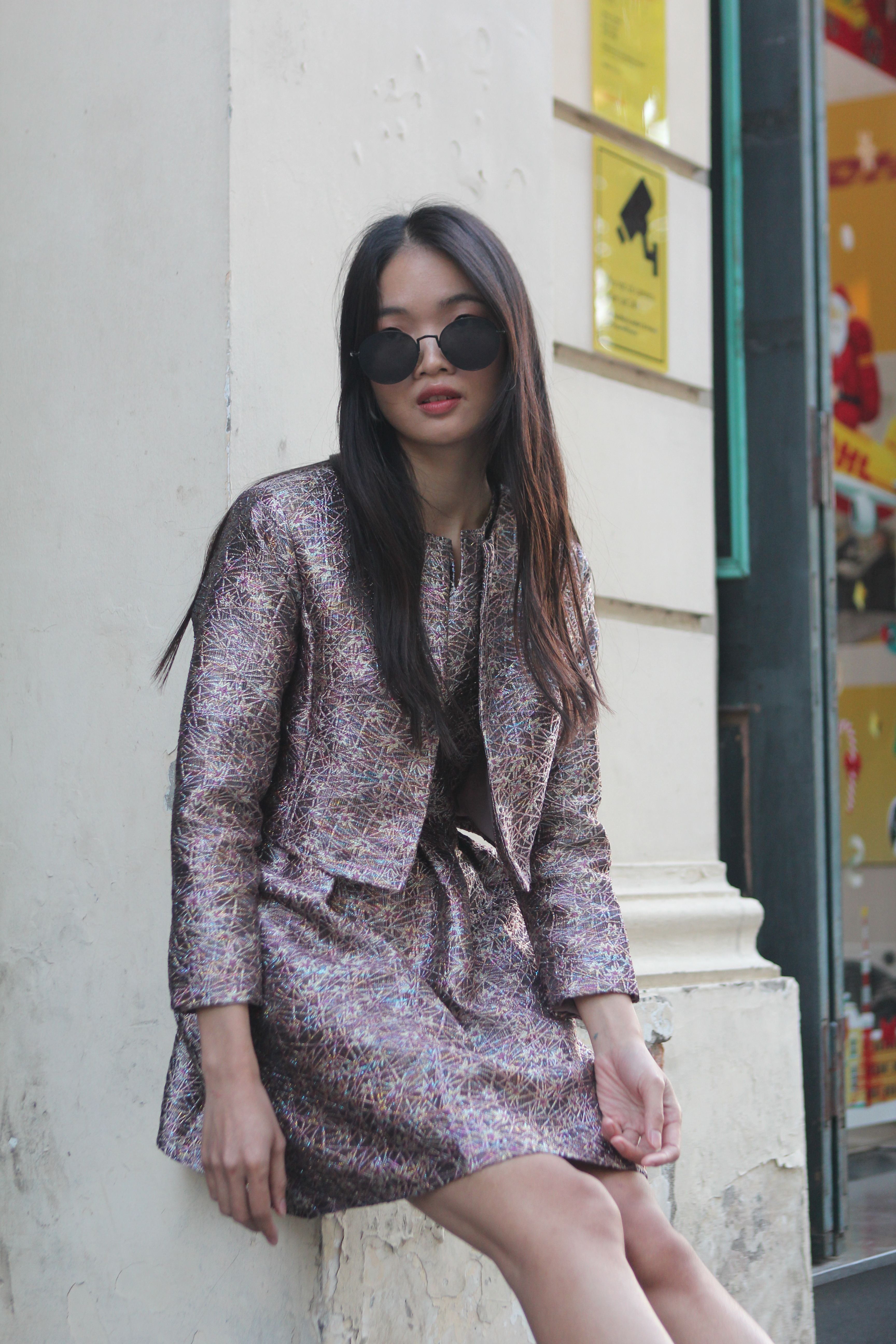 Fashionable woman in chic outfit and sunglasses sitting outdoors in city.